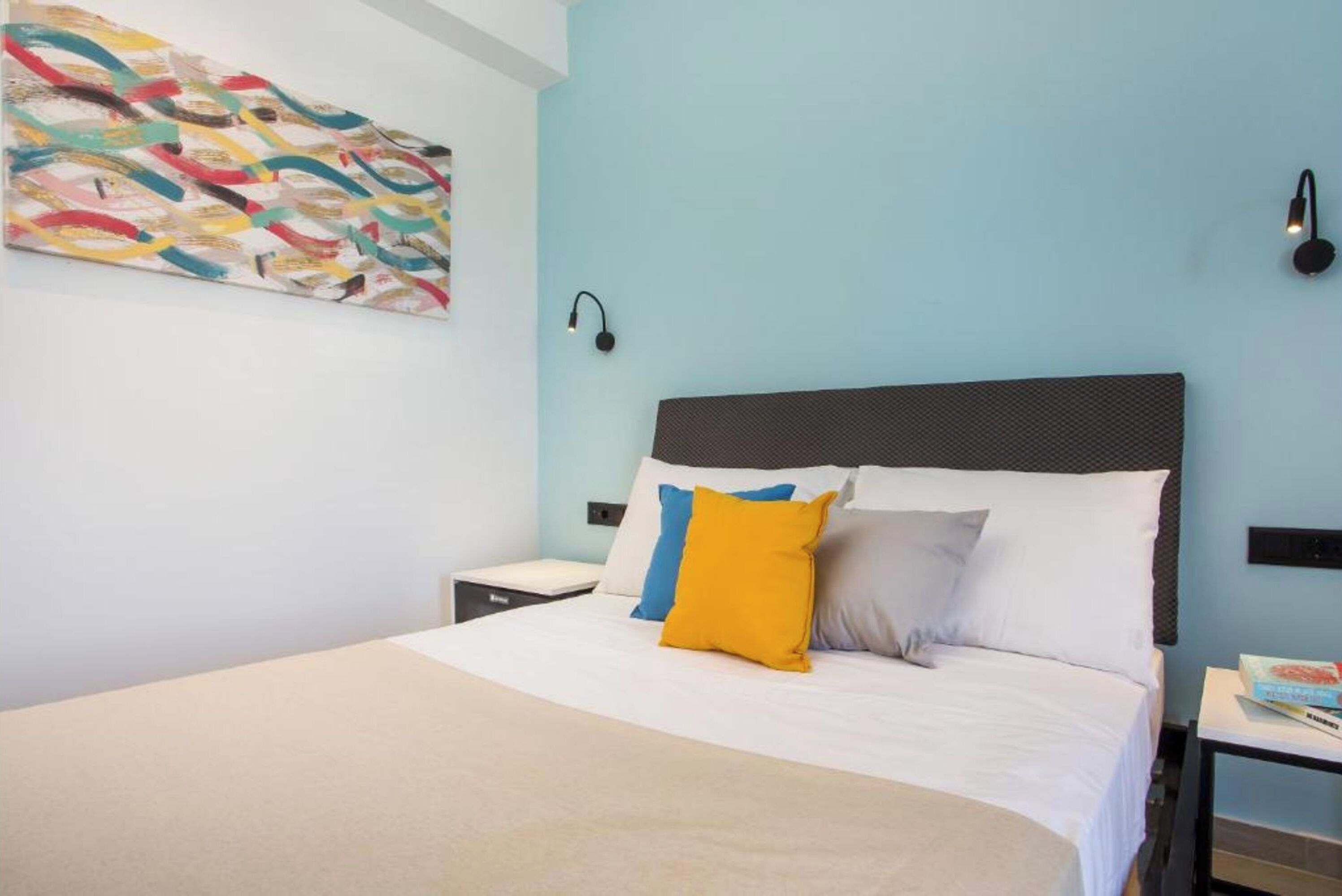 A neatly made bed with a white comforter and multiple pillows, a wall-mounted abstract painting, and a small bedside table with a lamp.