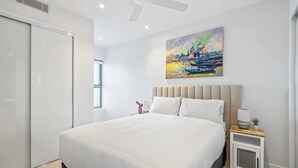 Un dormitorio moderno con una cama grande, un ventilador de techo, un cuadro de un paisaje urbano, una mesita de noche con una lámpara y un mueble auxiliar con una planta.