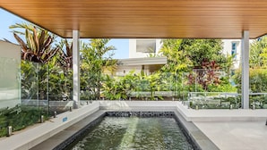 Zona de piscina exterior moderna con una pérgola de madera, rodeada de exuberante vegetación y barreras de vidrio.