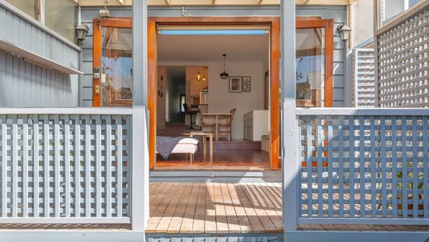 Um deck de madeira com cerca de lattic branco, uma porta de madeira e vista para um quarto com cama, mesa e cadeira.