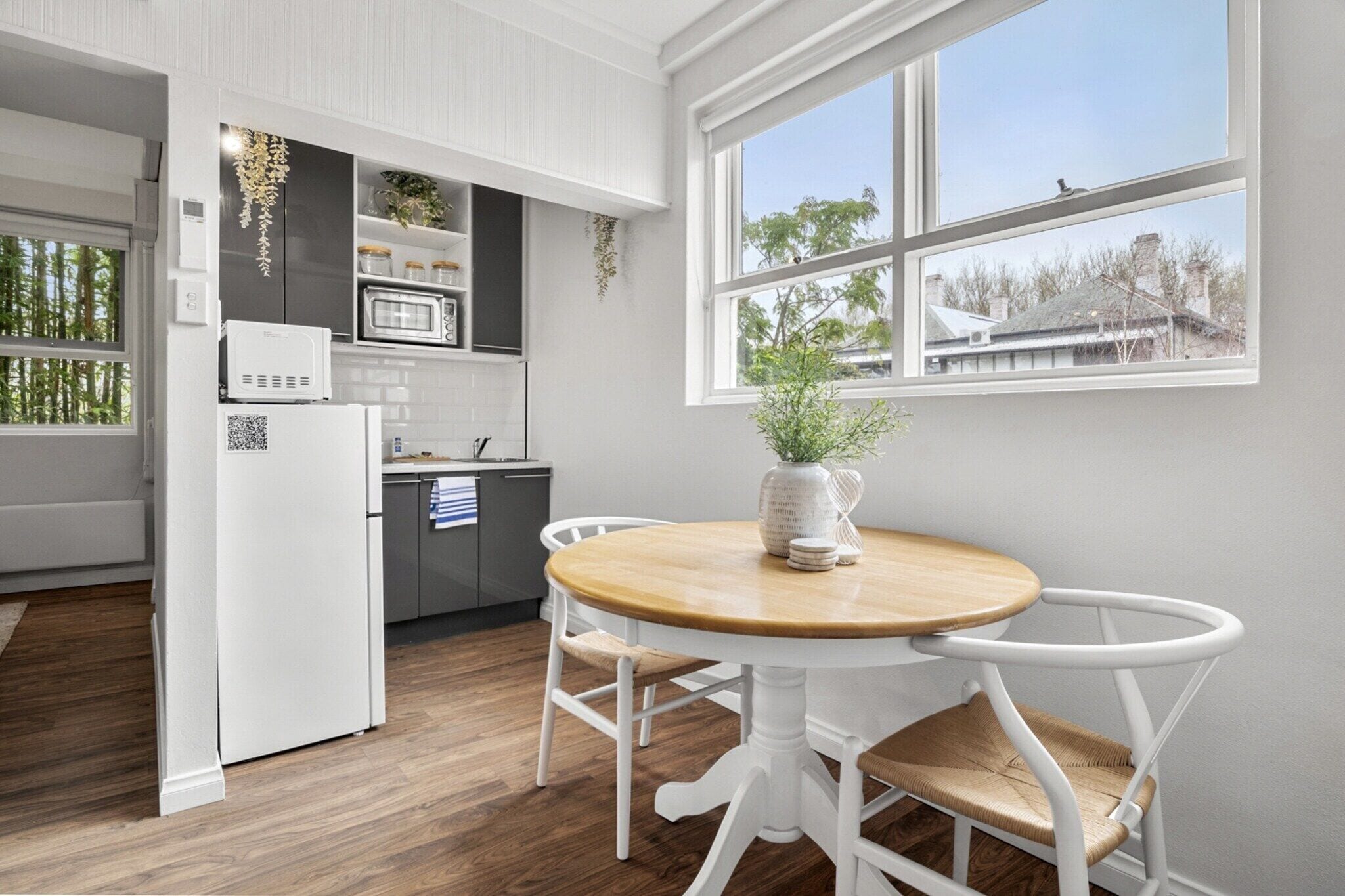 A modern kitchen with white cabinetry, a microwave, and a refrigerator. There is a dining table with white chairs and a vase with a plant.