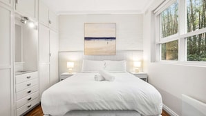 Une chambre à coucher avec un grand lit, des murs blancs, un tableau au-dessus du lit et une fenêtre donnant sur des arbres.
