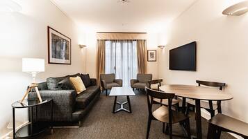 A hotel room with a sofa, two armchairs, a small table, a dining table with chairs, a flat-screen TV, and a large window with curtains.