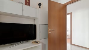 Een kamer met een televisie op een witte standaard, een houten deur en een witte muur.
