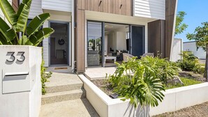 Una casa moderna de dos pisos con un amplio acceso por puerta de vidrio, un espacio habitable visible con sofá y mesa de centro, y un jardín bien cuidado con diversas plantas.