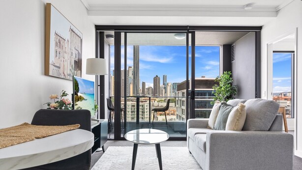 A modern living room with a sofa, a round table, and a view of a cityscape through large windows. - Chic 1 Bed Inner-City Retreat with Pool and Views (Fortitude Valley)