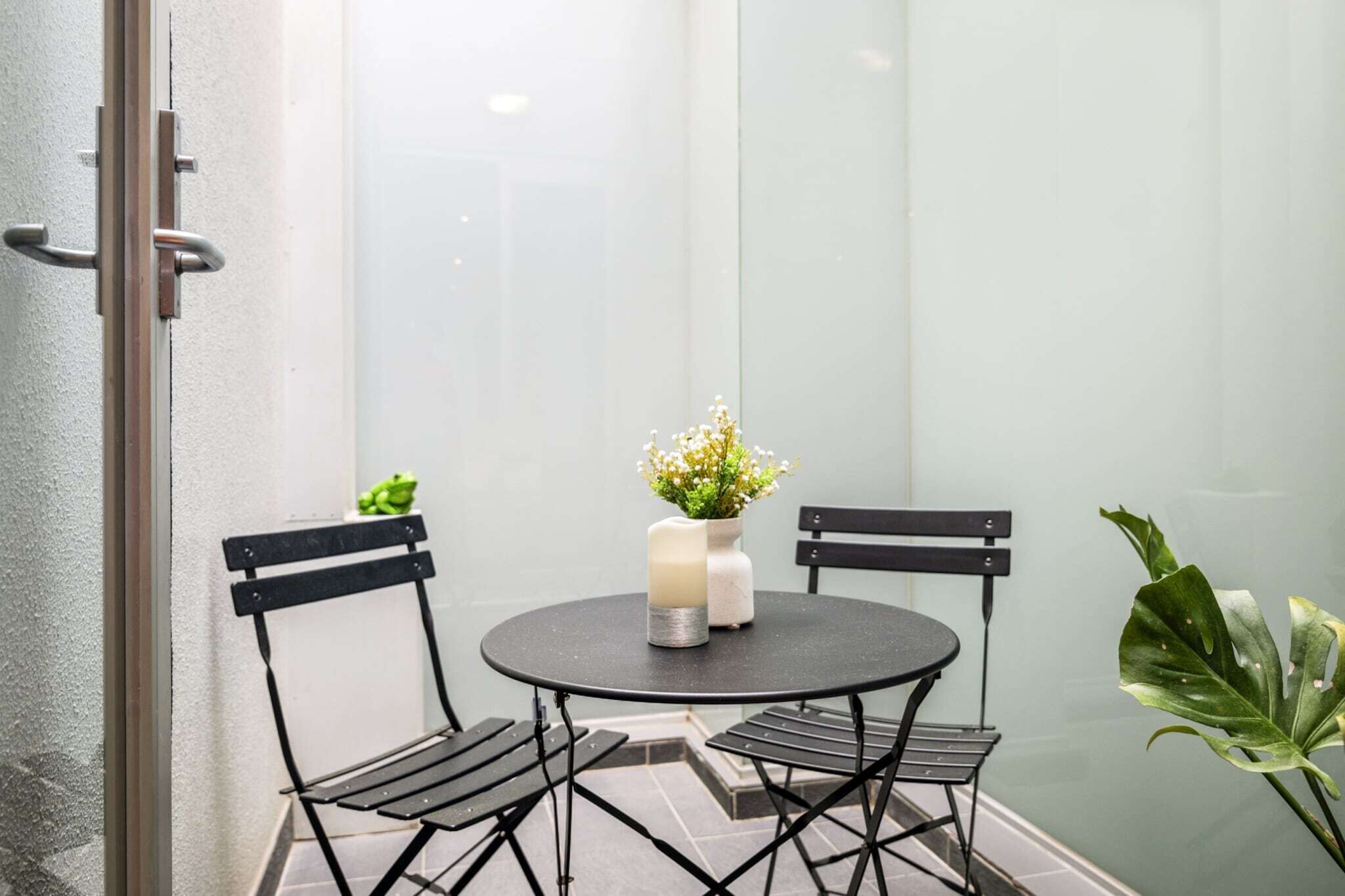 A small, enclosed patio area with a round table and two chairs, a vase with flowers, and a potted plant.