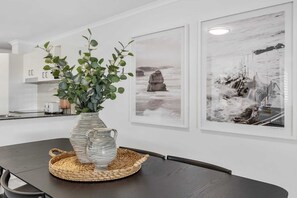Ein Essbereich mit einem dunklen Holztisch, einer gestreiften Vase mit Grünpflanzen und zwei gerahmten Schwarz-Weiß-Fotografien von Küstenlandschaften an der Wand.