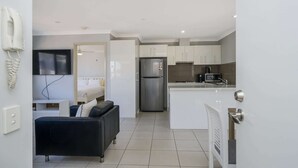 A modern kitchen with white cabinets, a stainless steel refrigerator, and a microwave. There is a black leather sofa and a white bedside table in the adjacent living area.