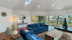 Uma sala de estar moderna com um sofá azul, uma mesa de centro de madeira e uma janela grande com vista para o verde.