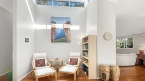 A spacious room with wooden flooring, two white cushioned chairs, a small round table, a bookshelf, and a large painting of a canyon. - Riverside 3-Bed Townhouse with Spacious Deck & BBQ (Mossy Point)