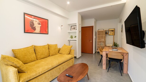 Ein Wohnzimmer mit einem gelben Sofa, einem Holztisch und einem an der Wand befestigten Fernseher.