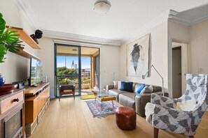Un soggiorno moderno con divano, poltrona e tavolino. È presente una zona cucina con un bancone e pensili. Un'ampia finestra offre vista sul verde.