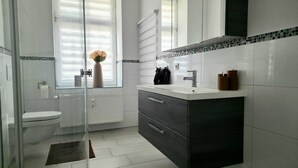 Une salle de bain moderne avec des toilettes, un lavabo surmonté d’un miroir et un vase de fleurs placé sur le rebord de la fenêtre.