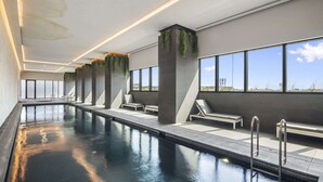 A modern indoor swimming pool with clear glass walls, lounge chairs, and a view of the landscape. - High-Rise 3-Bed Apartment with Premium Amenities (Box Hill)