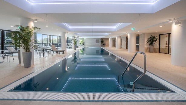 Ein modernes Hallenbad mit klarem Wasser, umgeben von Liegestühlen und Topfpflanzen, mit einer Decke mit Einbaustrahlern.