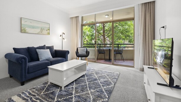 A modern living room with a sofa, coffee table, and TV. It features a large window with a view of greenery and a balcony. - Chic Urban Escape in Vibrant Camperdown (Camperdown)