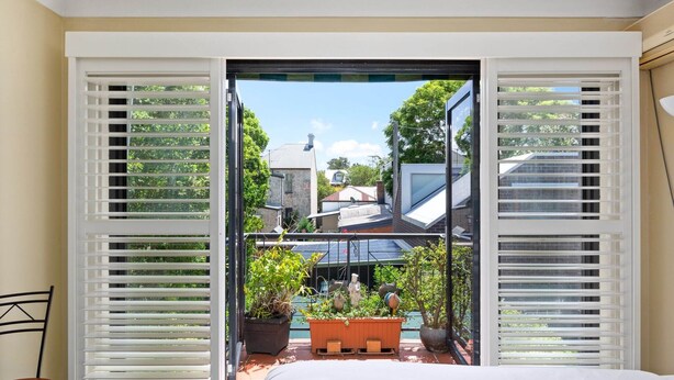 Een kamer met uitzicht op een tuin, zichtbaar door open witte shutters, een bed en een stoel.