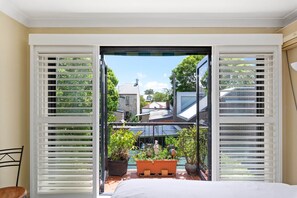 Une chambre donnant sur un jardin, avec des volets blancs ouverts, un lit et une chaise.