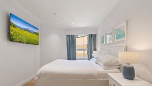 Ein Schlafzimmer mit Bett, Nachttisch mit Lampe, Fernseher an der Wand und Fenster mit Vorhängen.