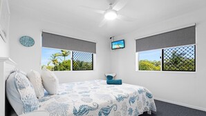 Ein Schlafzimmer mit Bett, Deckenventilator, Fernseher und zwei Fenstern mit Blick ins Grüne.