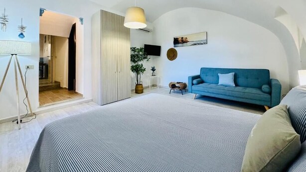 Ein modernes Schlafzimmer mit einem großen Bett, einem blauen Sofa und einem kleinen Tisch. Eine Tür führt in einen weiteren Raum, auf der Fensterbank steht eine Pflanze.