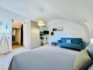 Ein modernes Schlafzimmer mit einem großen Bett, einem blauen Sofa und einem kleinen Tisch. Eine Tür führt in einen weiteren Raum, auf der Fensterbank steht eine Pflanze.