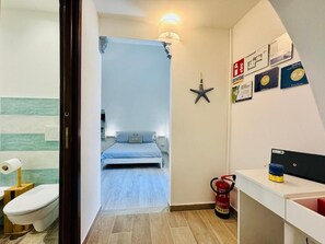 Ein schmaler Flur führt zu einem Schlafzimmer mit einem Bett und einem Nachttisch. Ein Feuerlöscher ist an der Wand angebracht.