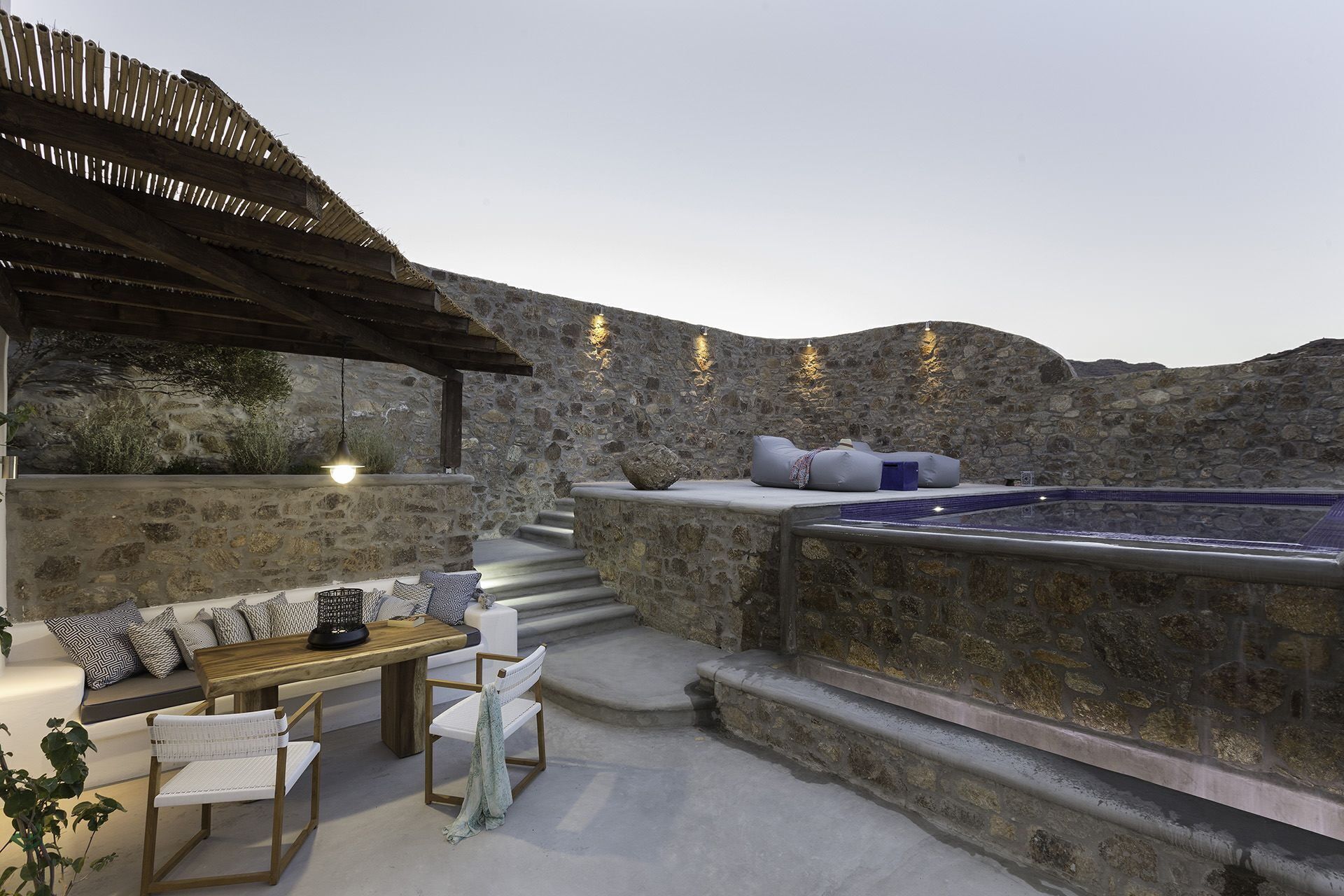 A stone-walled outdoor seating area with a pool, wooden furniture, and a pergola.