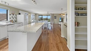 A modern kitchen with white cabinetry, a marble countertop, and a dining area with a table and chairs. - Coastal Upper-Level Home with Pool Access (Batehaven)