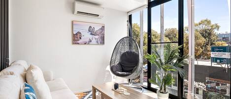 Una sala moderna con un sofá blanco, una mesa de centro de madera, un sillón colgante y un ventanal con vistas a la ciudad.