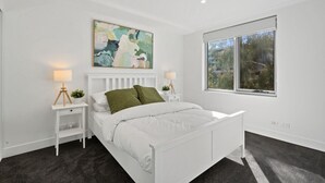 A modern bedroom with a white bed, two bedside tables with lamps, a large abstract painting, and a window with a view of trees. - Stylish 1-Bed Apartment with Balcony & Pool Access (Turner)