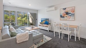 A modern living room with a grey sofa, a dining table with chairs, a TV, and a large window offering a view of trees and buildings. - Stylish 1-Bed Apartment with Balcony & Pool Access (Turner)