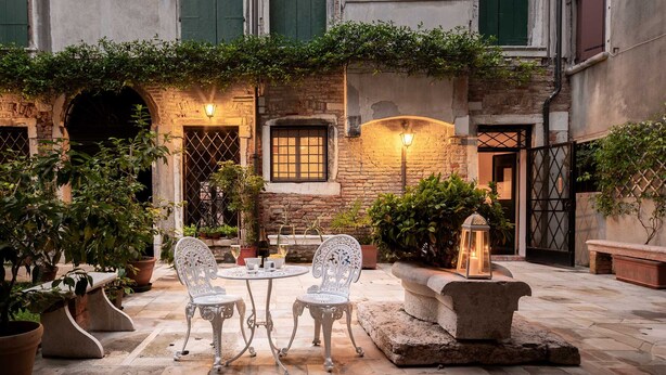 Une terrasse extérieure avec un sol en pierre, deux chaises en métal blanc, une petite table ronde et des plantes en pot.