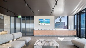 Un área de espera moderna con sofás grises, una mesa de centro de mármol, un mostrador de recepción de madera y un amplio ventanal con vista a la playa.