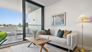 A modern living room with a sofa, coffee table, and floor lamp. A large window offers a view of the outdoors. - Modern 1-Bed Apartment with Views in Fremantle (North Fremantle)