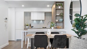 A modern kitchen with white cabinetry, a wooden dining table, and black chairs. There is a plant on the right side and a shelf with decorative items. - Charming 1-Bed with Rooftop Access & Parking (North Fremantle)