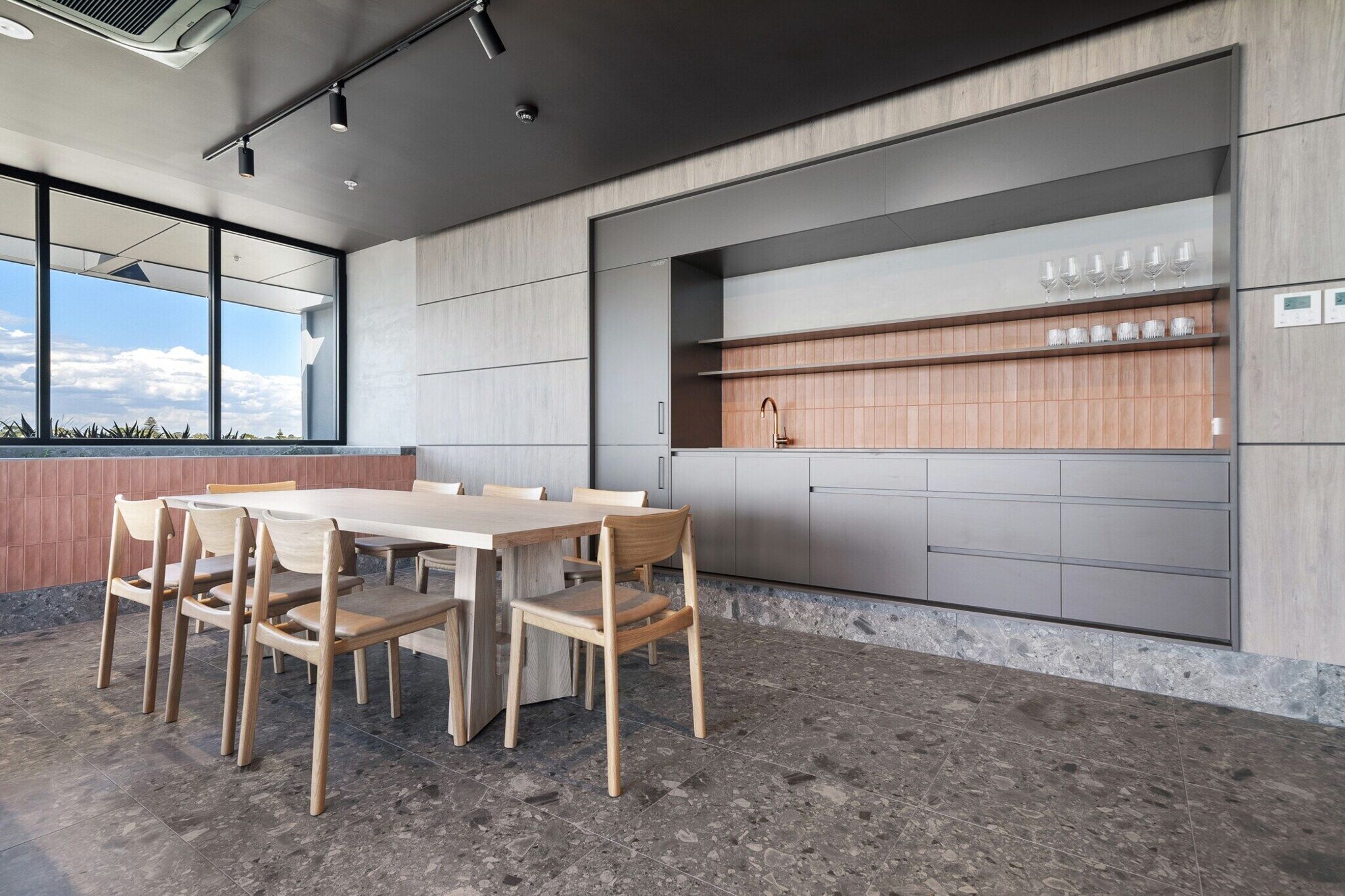 A modern kitchen with a built-in oven, a wooden dining table with chairs, and a large window offering a view of the sky.