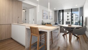 Et moderne køkken med en central ø, et spiseområde med et rundt bord og stole, samt store vinduer med udsigt over byen.