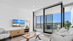 A modern living room with a large window, a flat-screen TV, a wooden coffee table, and a sofa. - Stylish 2-Bed Unit with Views & Rooftop Access (North Fremantle)