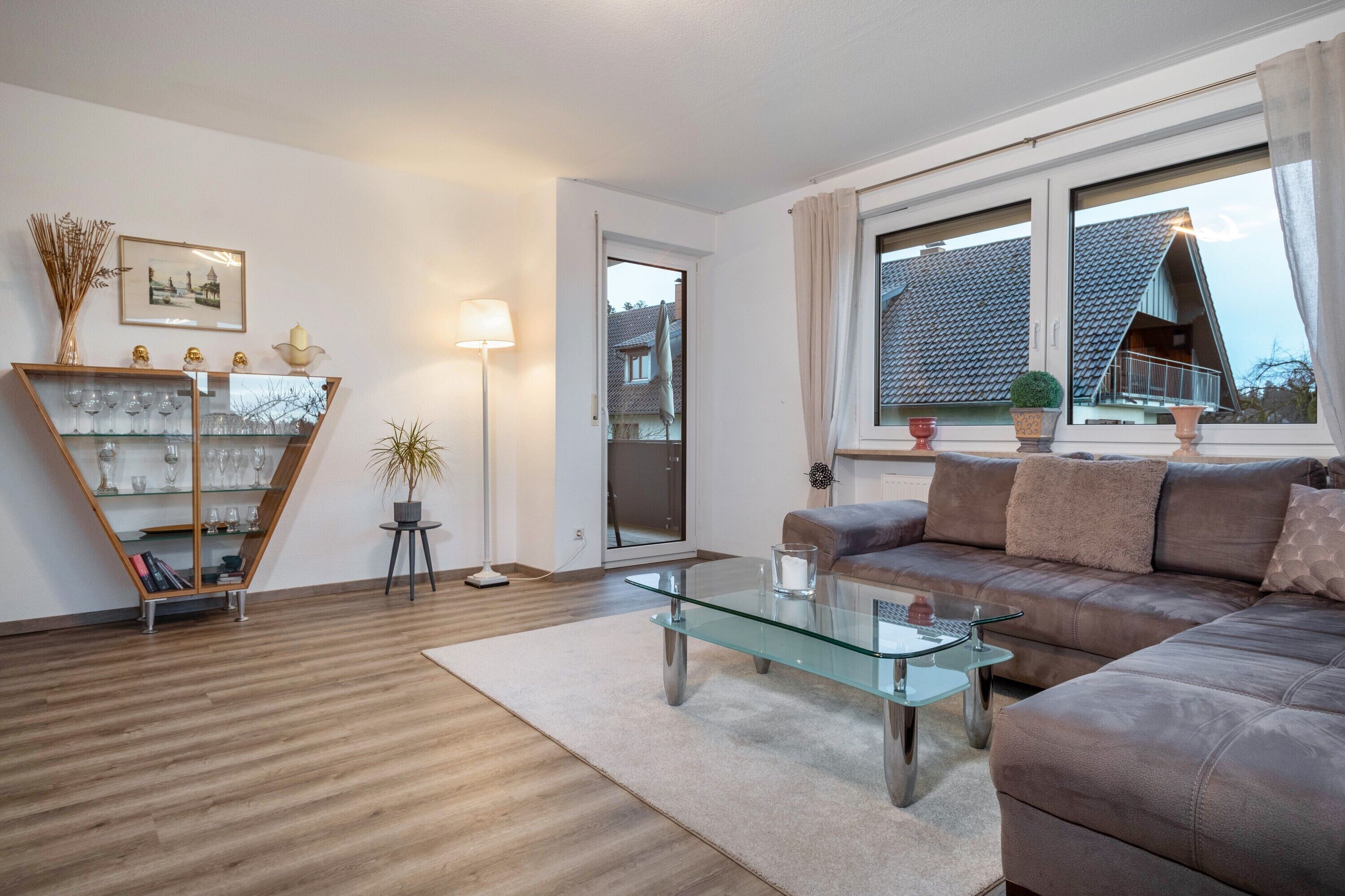 Ein modernes Wohnzimmer mit einem Glastisch, einem braunen Ledersofa, einer Glastrennwand und einem großen Fenster mit Blick auf ein Haus.