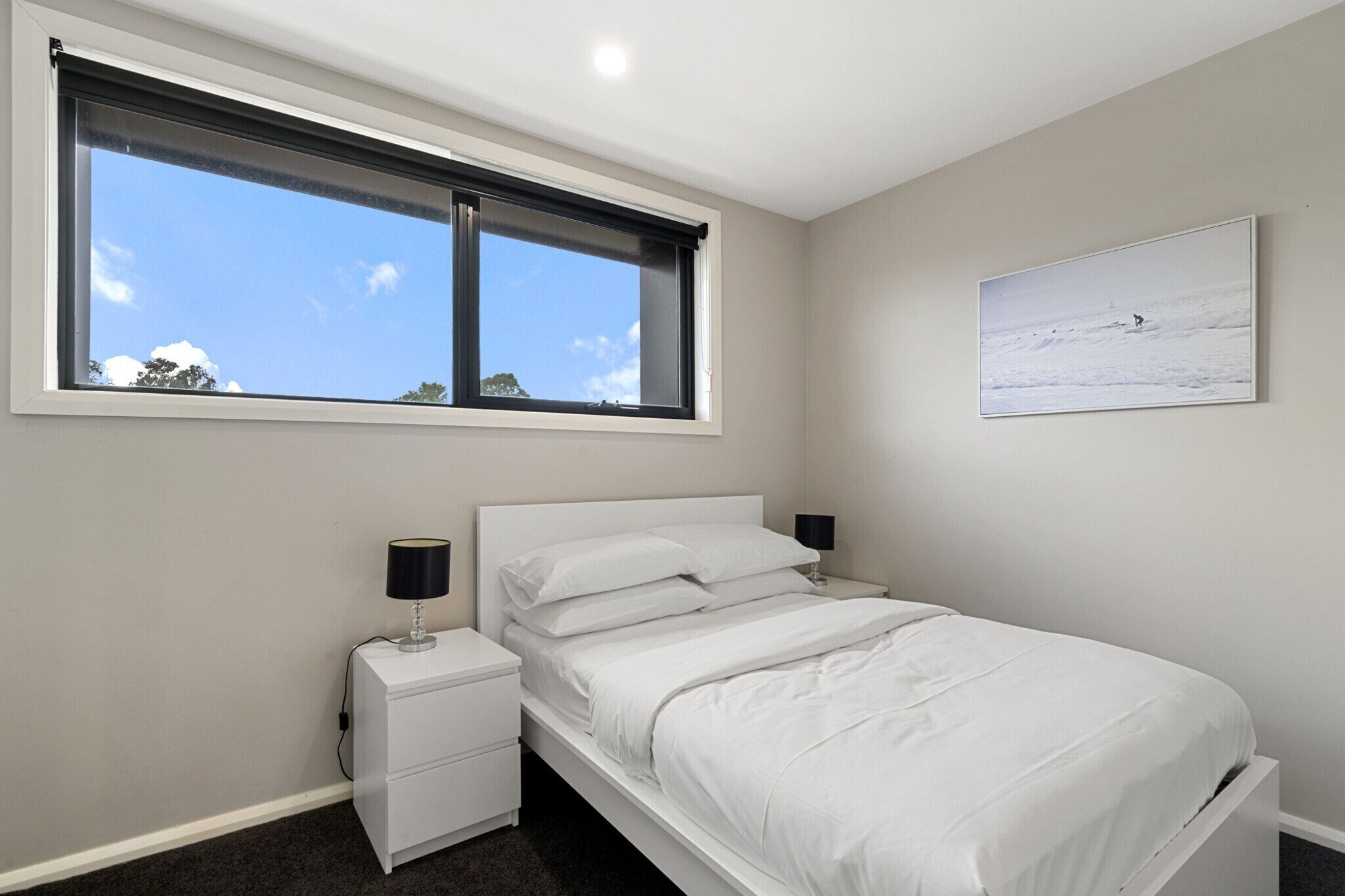A bedroom with a bed, a nightstand, a lamp, a window with blinds, and a painting on the wall.