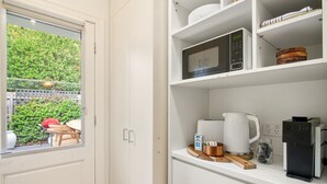 Cuina amb armaris blancs, microones, cafetera i finestra amb vista a un jardí.