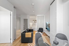 Ein modernes Wohnzimmer mit einer Couch, einem Couchtisch und einem Fernseher. Eine Glastrennwand und eine Tür führen in einen anderen Raum.
