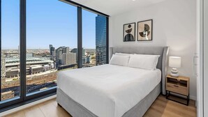 Une chambre d’hôtel moderne dotée d’un grand lit, d’une table de chevet et offrant une vue sur le paysage urbain grâce à de grandes fenêtres du sol au plafond.