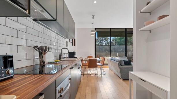 Una cocina moderna con baldosas blancas, encimeras de madera y electrodomésticos de acero inoxidable. Cuenta con una zona de comedor con una mesa y sillas, y un sofá gris al fondo.