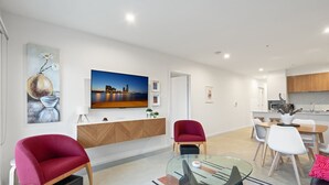 Un salon moderne avec une table basse en verre, des fauteuils rouges et blancs, un téléviseur à écran plat et un tableau représentant un paysage urbain.