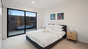 Une chambre avec une grande porte-fenêtre coulissante donnant sur un balcon, un lit avec une literie blanche, deux tableaux accrochés au mur et une table de chevet avec une lampe.