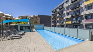 A swimming pool with lounge chairs and a blue canopy, surrounded by multi-story buildings with balconies. - Central East Perth Flat with Pool & Gym Access (East Perth)