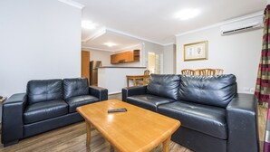 A living room with two black leather sofas, a wooden coffee table, and a dining area with a wooden table and chairs. - Central East Perth Flat with Pool & Gym Access (East Perth)